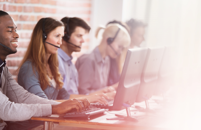 A diverse group of call center representatives wearing headphones with microphones in line at a desk working on their computers.