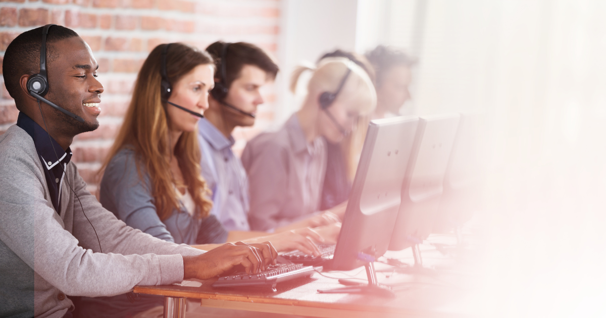 A diverse group of call center representatives wearing headphones with microphones in line at a desk working on their computers.