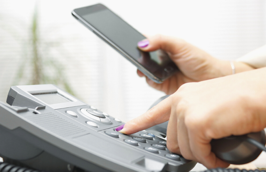 A woman holding a business landline telephone and dialing a phone number with one hand and holding a smartphone in the other hand.