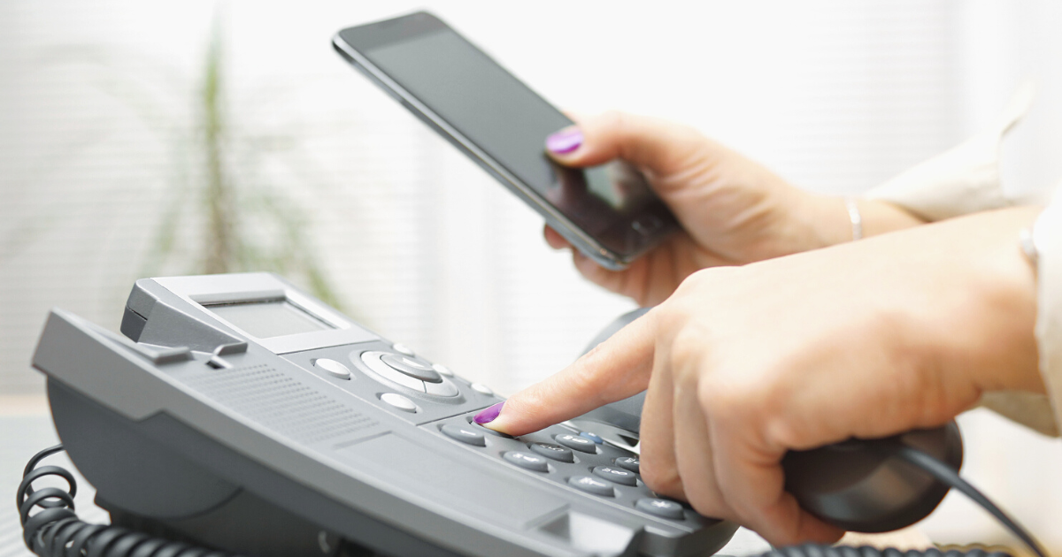 A woman holding a business landline telephone and dialing a phone number with one hand and holding a smartphone in the other hand.